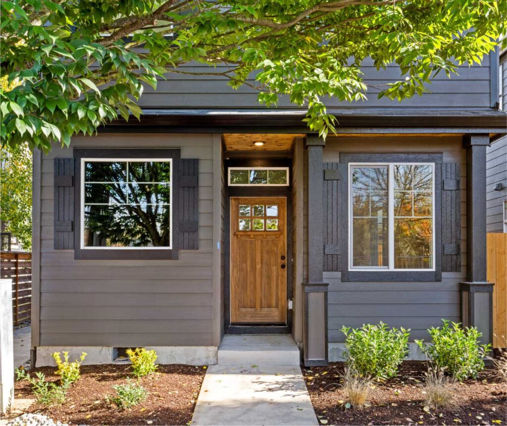 Front entrance of a gray house with a wooden door, two windows with dark shutters, and a small landscaped garden. References a tree canopy above.