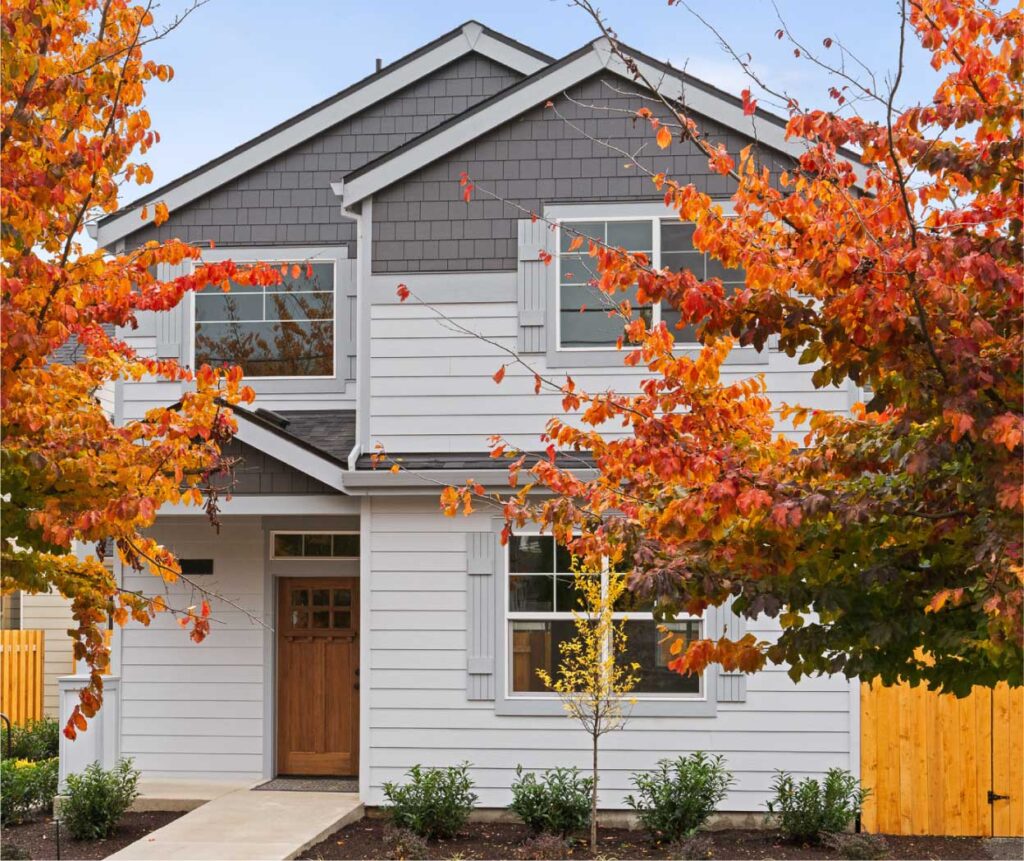 Two-story suburban house with gray siding and a wooden front door, framed by vibrant orange autumn leaves in the foreground.