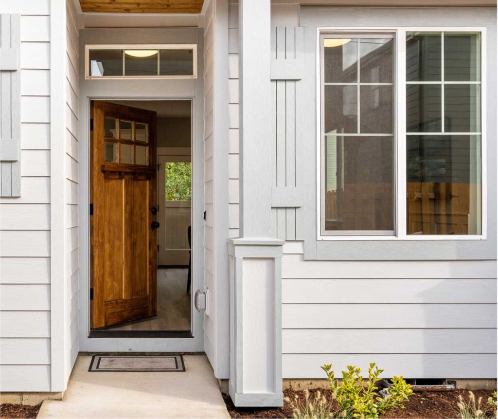 Open wooden front door of a light-gray house, showing a small entry porch and a large window to the right.
